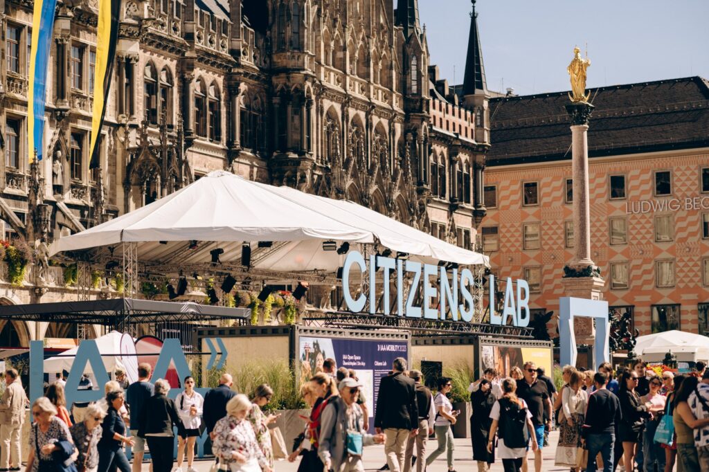 Das Bild zeigt eine belebte Szene vor historischen Gebäuden, vermutlich auf einem Marktplatz oder einem öffentlichen Platz in einer Stadt. Im Vordergrund sind viele Menschen zu sehen, die dort flanieren oder sich versammeln. Im Mittelpunkt steht ein großes Schild mit der Aufschrift „CITIZENS LAB“, das auf eine Veranstaltung oder ein Projekt hinweist. Dahinter ist ein großes Zelt aufgebaut, vermutlich für eine Ausstellung oder ein Event. Die Atmosphäre wirkt lebendig und urban, mit einer Mischung aus moderner Aktivität und historischem Flair.