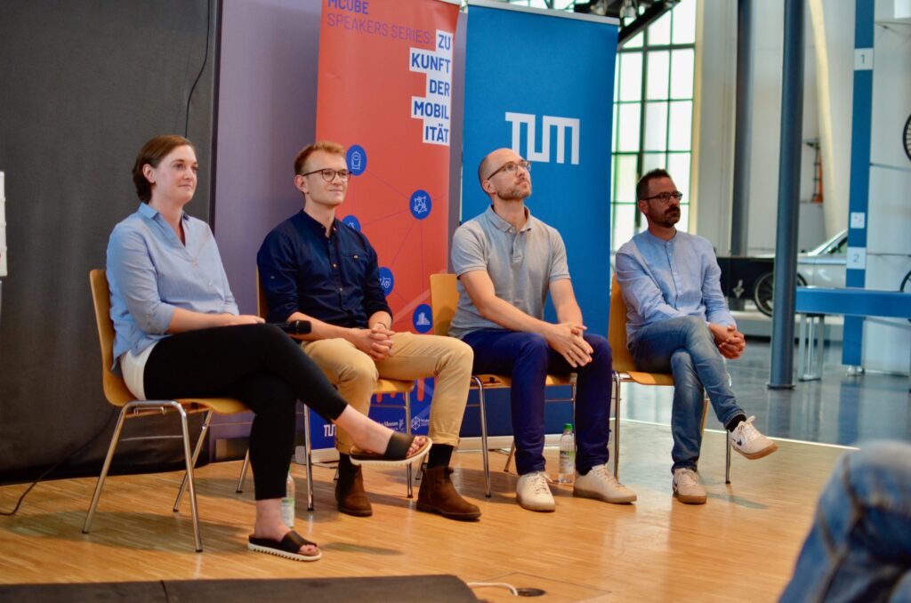 Das Bild zeigt eine Podiumsdiskussion mit vier Personen, die auf Stühlen nebeneinander sitzen. Drei Männer und eine Frau sitzen entspannt und aufmerksam da, sie tragen legere Kleidung. Im Hintergrund sind Banner mit dem TUM-Logo und der Aufschrift „MCUBE SPEAKERS SERIES: ZUKUNFT DER MOBILITÄT“ zu sehen. Die Atmosphäre wirkt professionell und fokussiert, als ob eine Diskussion oder ein Vortrag zum Thema Mobilität stattfindet.