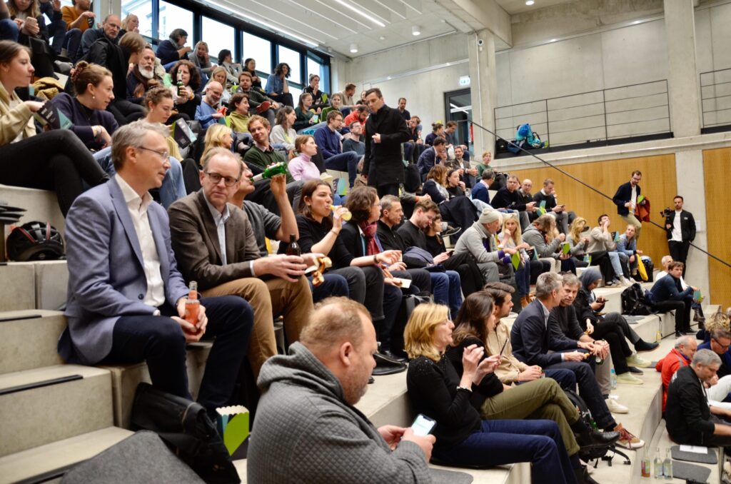 Ein gut besetztes Auditorium mit zahlreichen Menschen, die auf gestuften Sitzreihen Platz genommen haben. Einige Personen unterhalten sich, andere trinken oder nutzen ihr Smartphone. Die Atmosphäre wirkt lebendig und konzentriert.