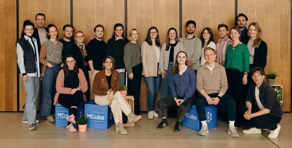 Auf dem Foto ist das Team von MCube Mosaiq zu sehen. Einige Personen sitzen auf blauen Würfeln mit der Aufschrift "MCube", andere stehen dahinter. Im Vordergrund befinden sich auch kleine grüne Pflanzen in Töpfen.