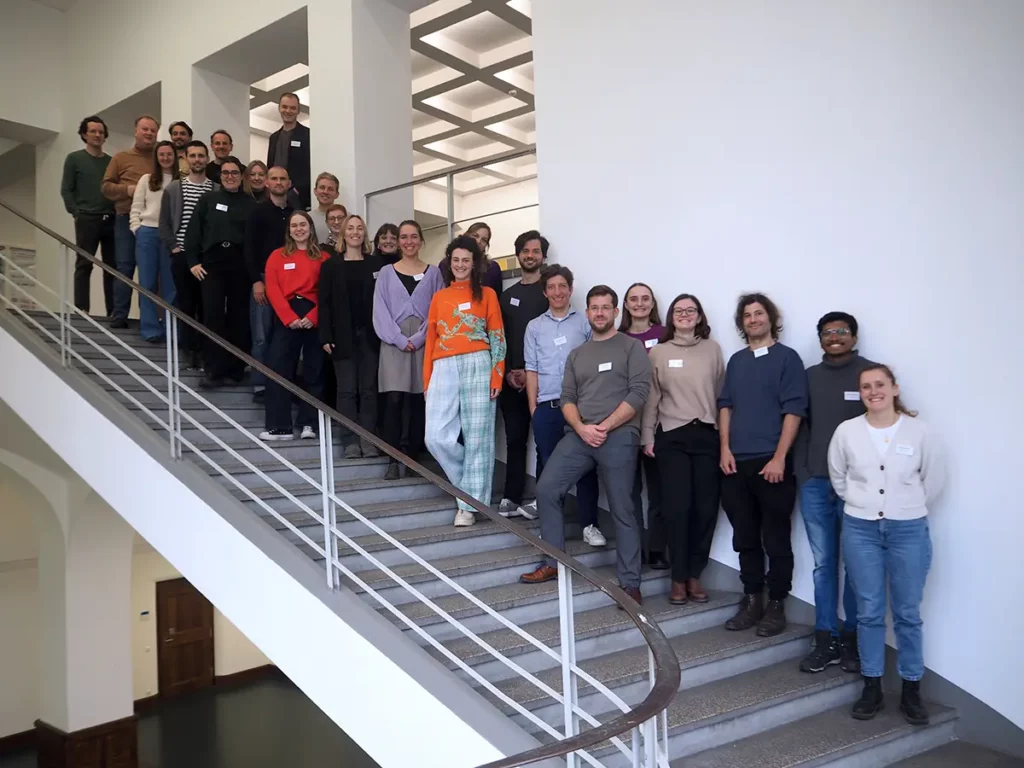 Gruppenfoto von etwa 30 Personen, die sich auf einer Treppe im Innenbereich aufstellen. Die Personen sind locker angeordnet und blicken in die Kamera. Die Umgebung wirkt modern mit weißen Wänden und einem großen Treppenaufgang mit Geländer. Die Gruppe besteht aus Männern und Frauen unterschiedlichen Alters, die lächelnd.