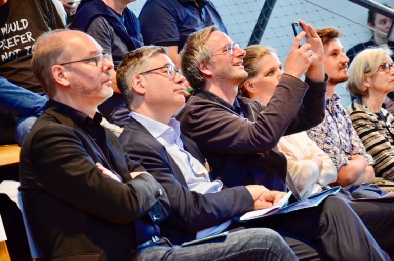 Blick auf die erste Reihe des Publikums von der Seite, während der MCube Speaker Series #20 mit dem Thema "Olympia als Entwicklungsmotor?", die interessiert dem Beginn der Veranstaltung lauschen.